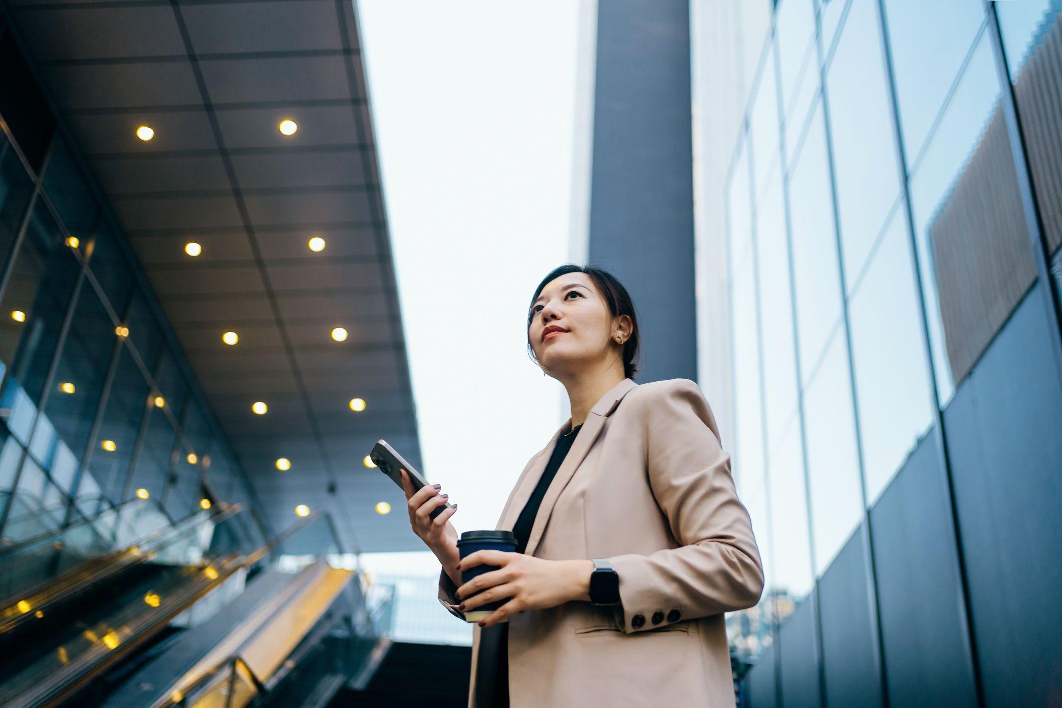A woman in a beige blazer holding a phone and coffee cup, looking up in a modern city setting.